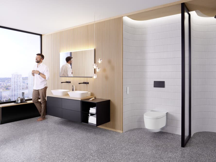 Bărbat într-o baie mare, cu mobilier închis la culoare și vas wc cu funcție de bideu Geberit AquaClean Bărbat într-o baie mare, cu mobilier închis la culoare și vas wc cu funcție de bideu Geberit AquaClean